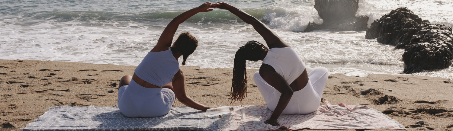 Two women in activewear performing a seated side stretch on the beach, facing the ocean, with waves crashing in the background.