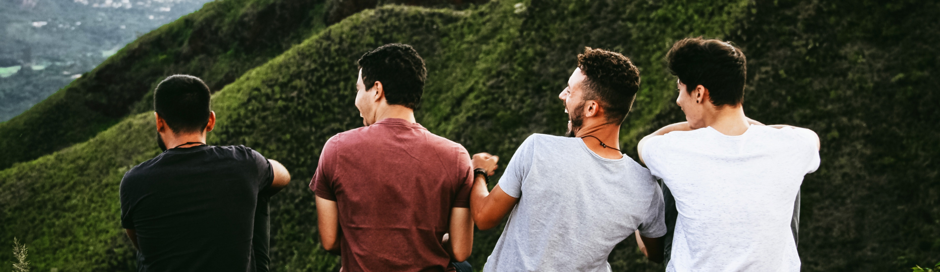 Group of four young men sitting outdoors on a hillside, enjoying nature and laughing together with lush green mountains in the background.