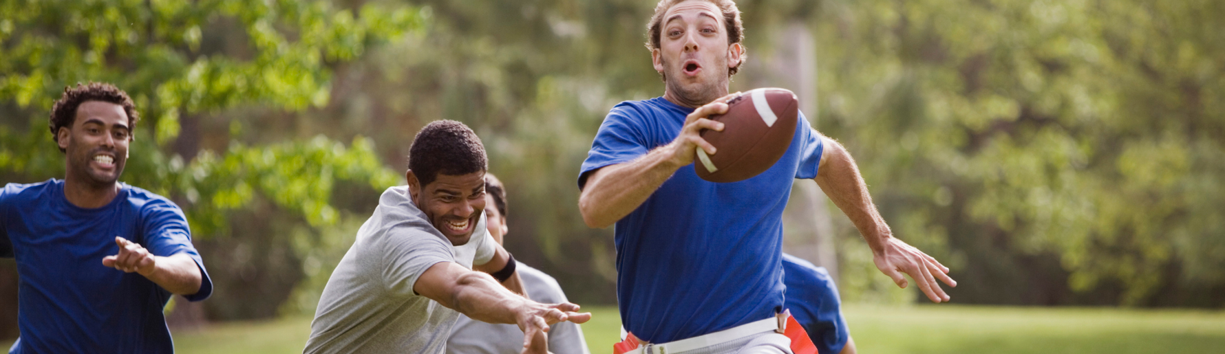 A group of men playing flag football outdoors in a park. One man, wearing a blue shirt, is running with the football while others, in gray shirts, are chasing him. The scene is set in a grassy area with trees in the background.
