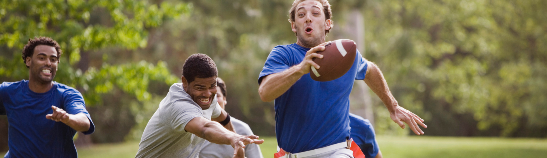 A group of men playing flag football outdoors in a park. One man, wearing a blue shirt, is running with the football while others, in gray shirts, are chasing him. The scene is set in a grassy area with trees in the background.