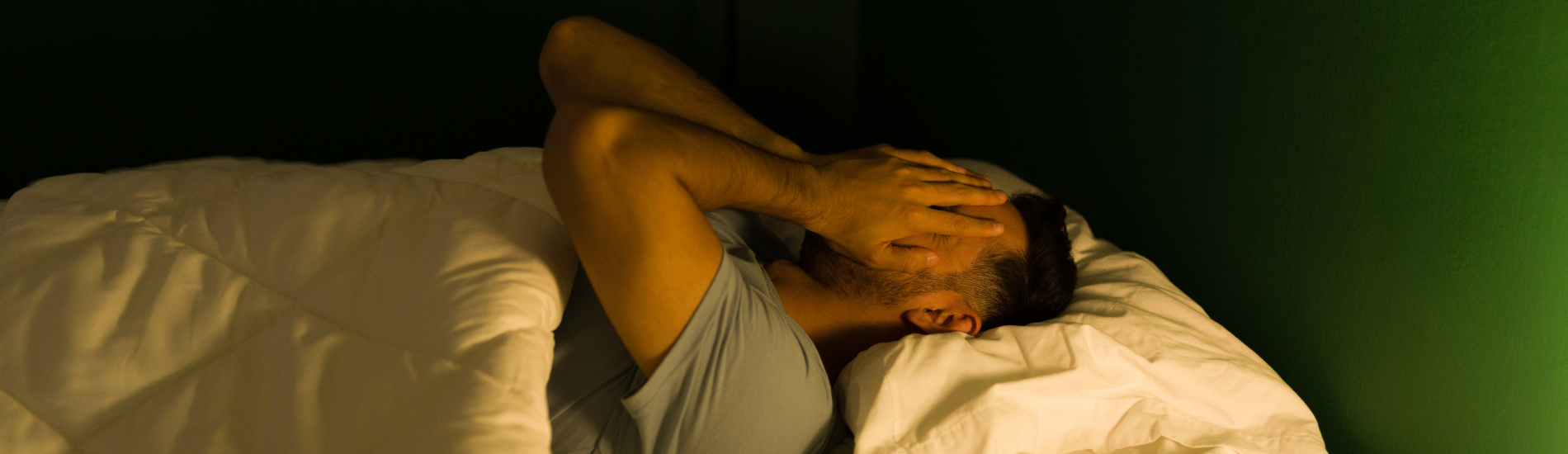 A man lies in bed, holding his head with both hands in frustration, possibly experiencing difficulty sleeping or discomfort.