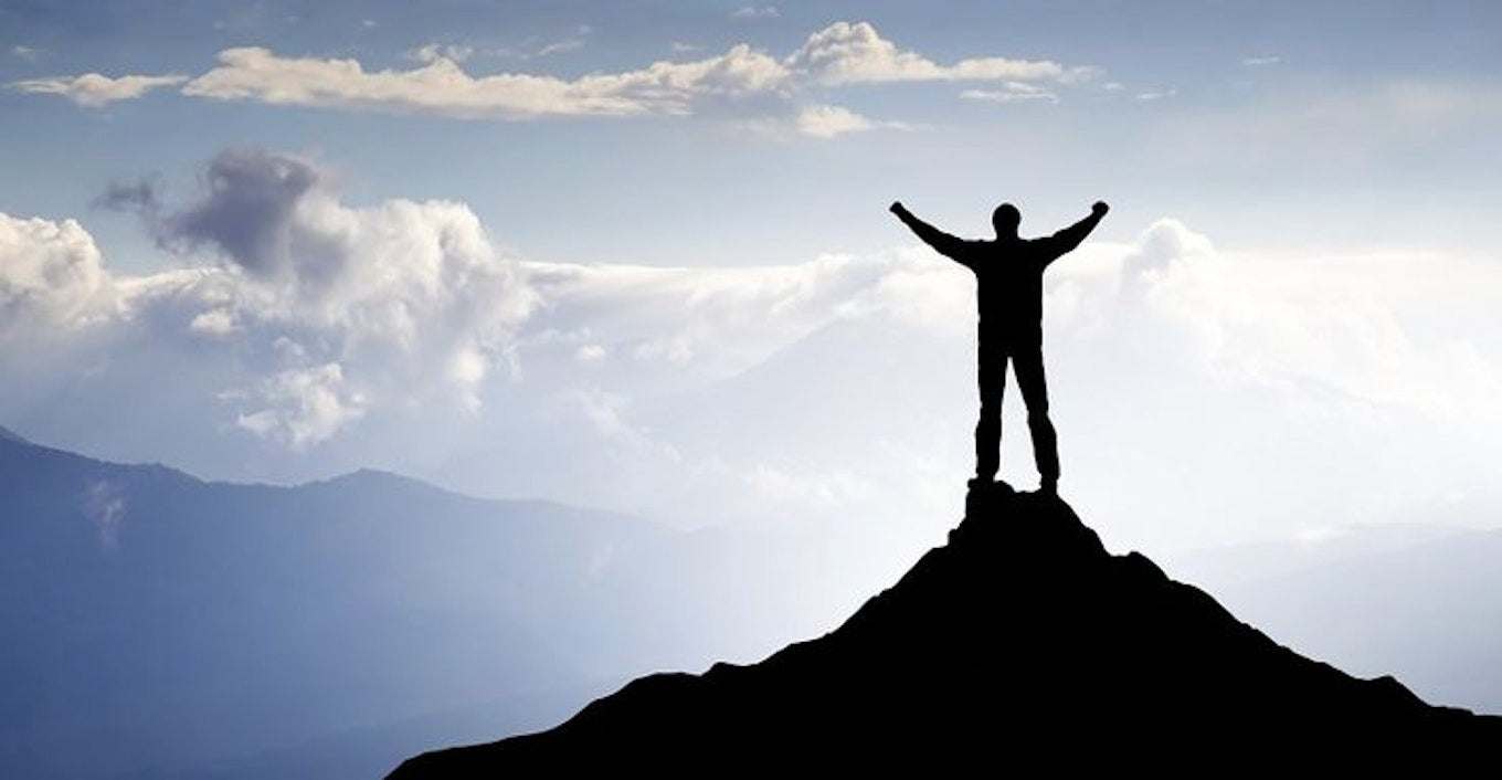 a person sitting at the top of a mountain with clouds and other mountains in the distance, the person’s hands are in the air, signifying the achievement of being at the top of the world